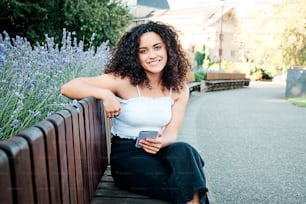 Attractive curly-haired woman sitting on a park chair looking at the camera and smiling holding her cell phone in her hand.