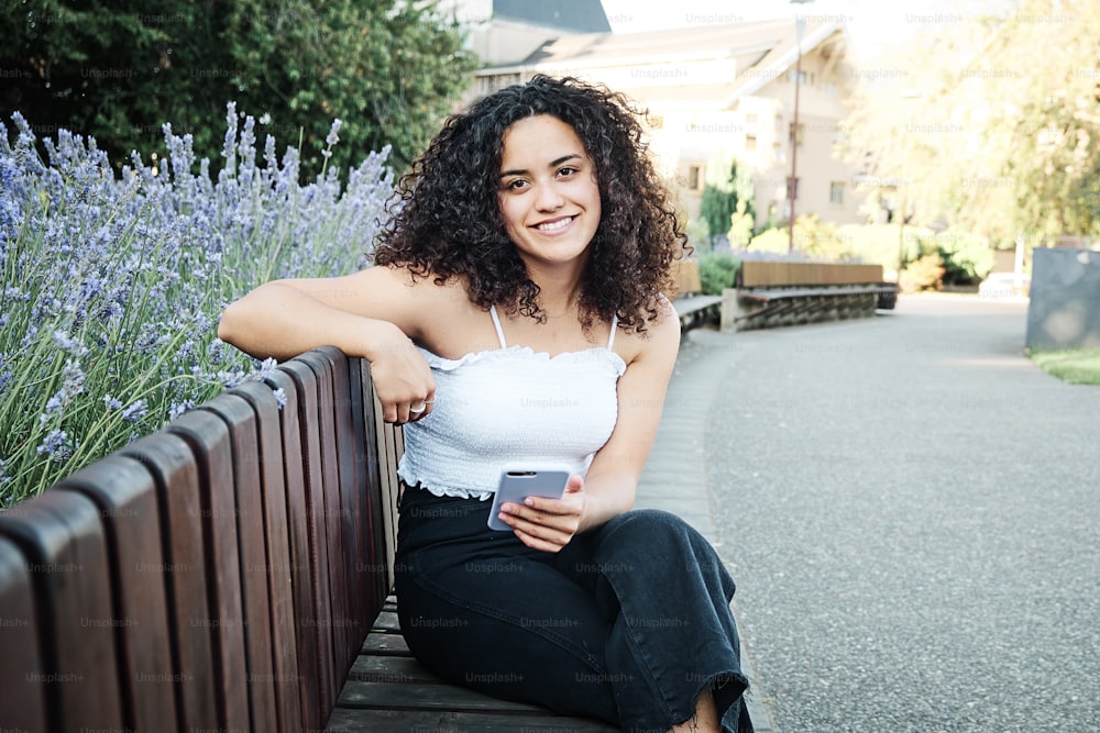 Attractive curly-haired woman sitting on a park chair looking at the camera and smiling holding her cell phone in her hand.