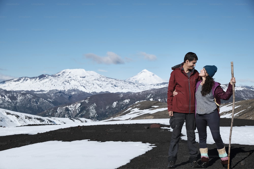Verliebtes Paar, das auf einem Felsen steht und sich ansieht und lächelt. Im Hintergrund blauer Himmel und schneebedeckte Berge, südlich von Chile.