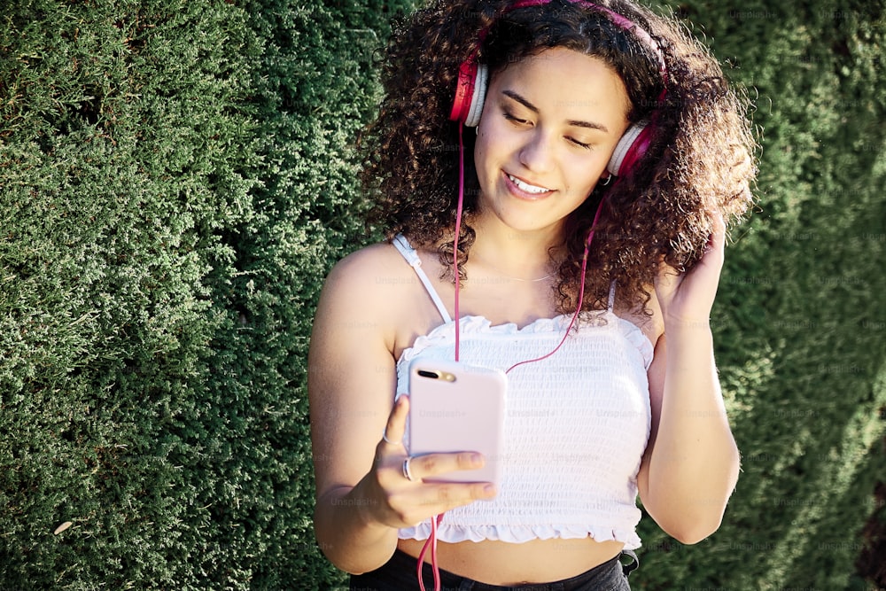 Young curly-haired woman is listening to music with headphones and looking at her cell phone leaning against a natural green wall.