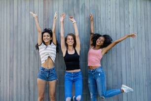 Three best female friends jumping together outdoors near a wall, looking at camera