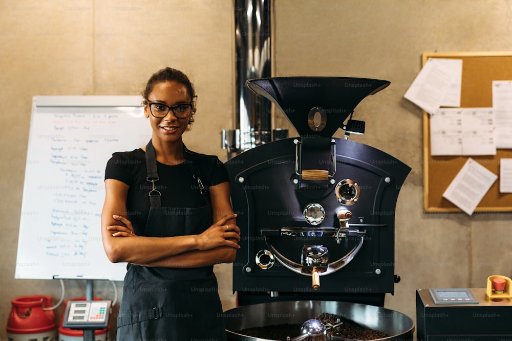 喫茶店のコーヒー焙煎機の横に立つ女性バリスタ。カメラを見つめる笑顔の経営者。