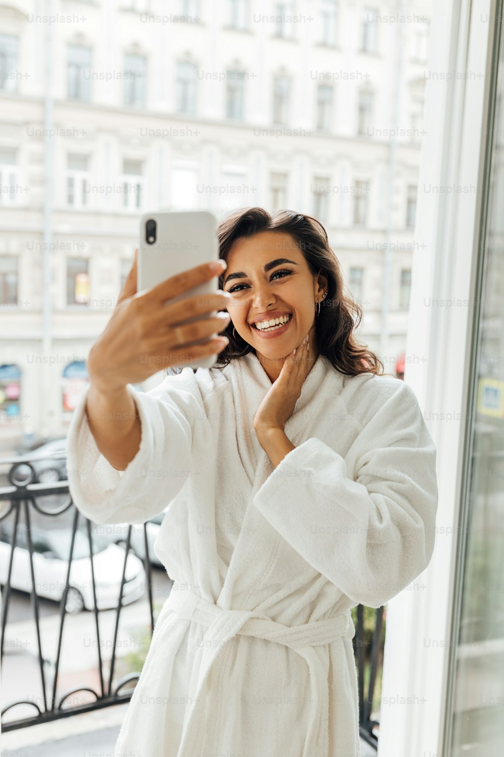Happy woman taking selfie on a balcony. Young female in bathrobe holding a smartphone and making photos.