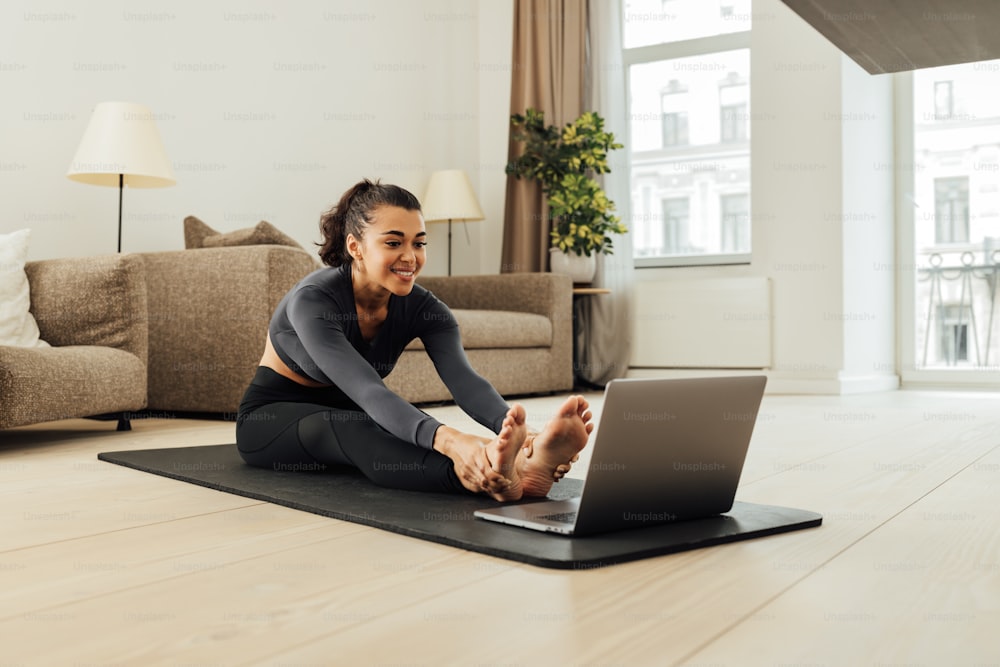 Smiling woman in sportswear sitting on a mat in front of a laptop and stretching her body