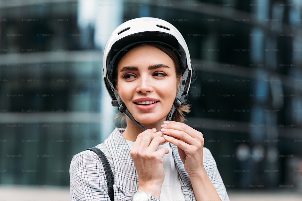 Donna d'affari sorridente che si allaccia un casco da ciclista mentre si trova in città. Giovane femmina che mette un casco bianco sulla sua testa.