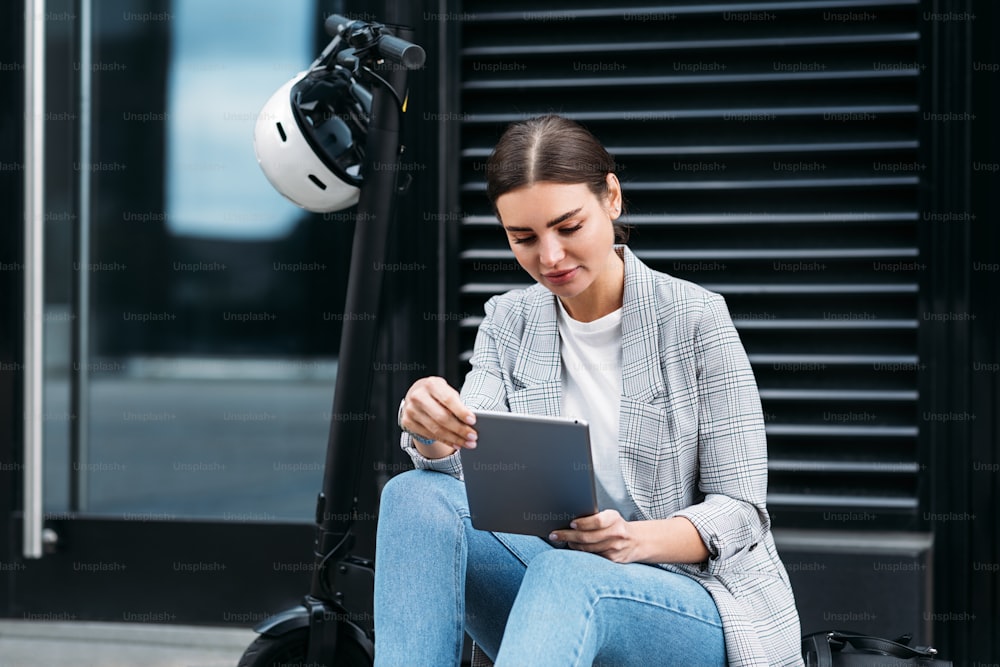 Schöne kaukasische Frau, die auf einem geparkten Elektroroller auf der Straße sitzt und ein digitales Tablet hält