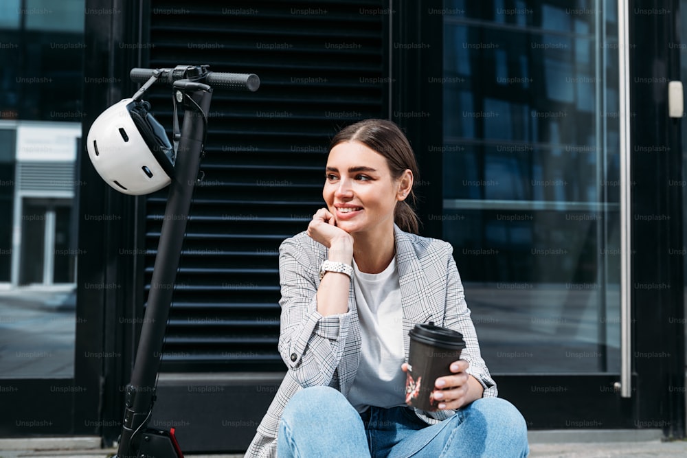 Belle femme d’affaires souriante avec un café assise sur un scooter électrique dans un immeuble regardant au loin