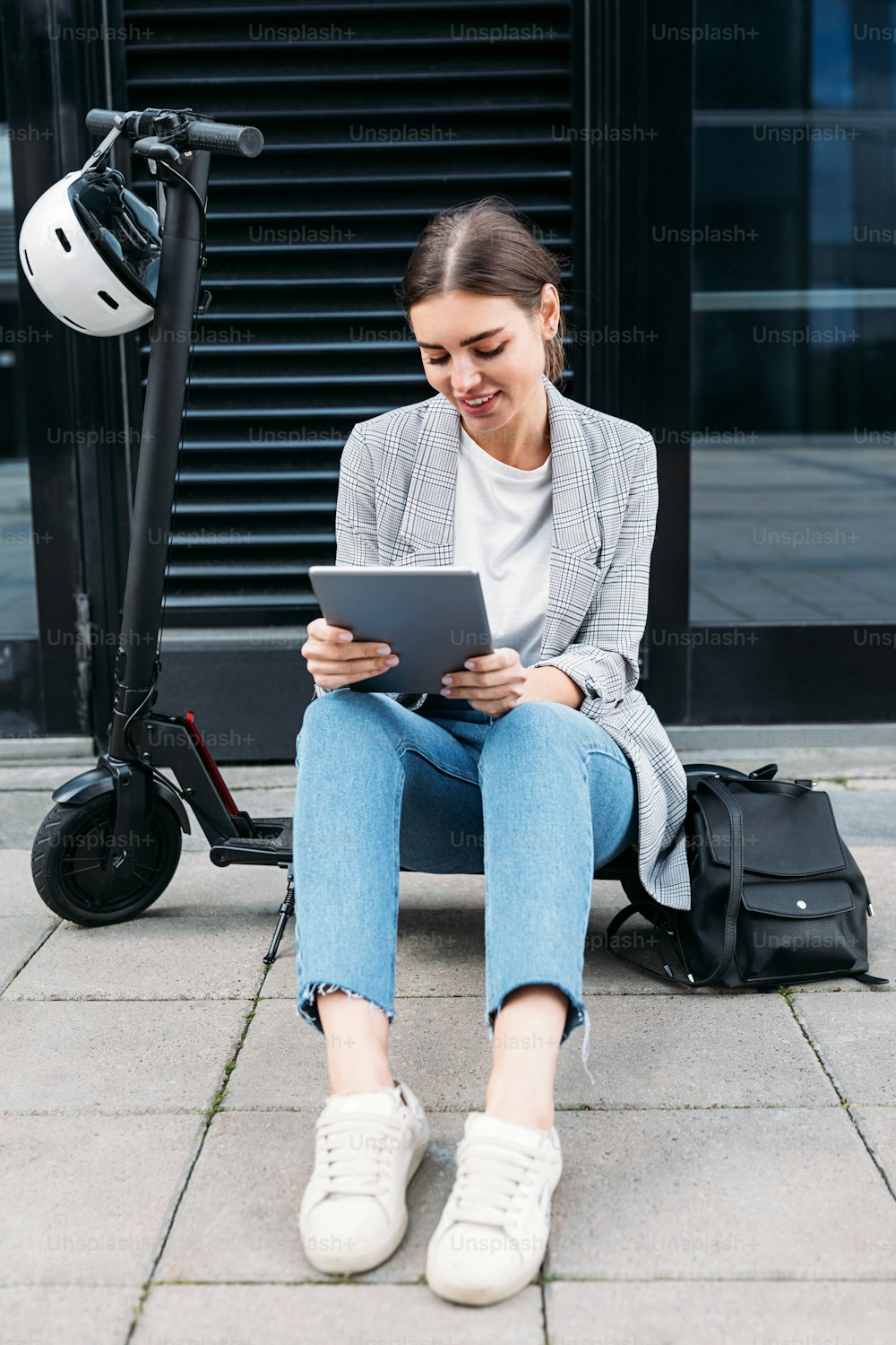 Mulher nova em casuais sentados em uma scooter elétrica no edifício segurando um tablet digital