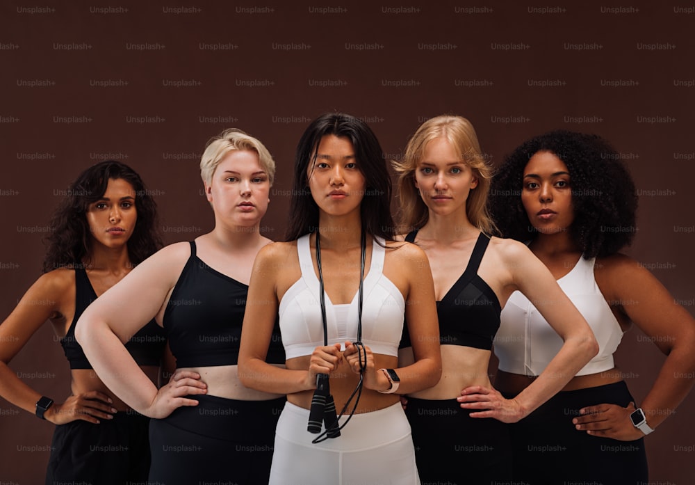 Five women in sportswear looking at camera. Females with different body types and ethnicities standing together against brown background.