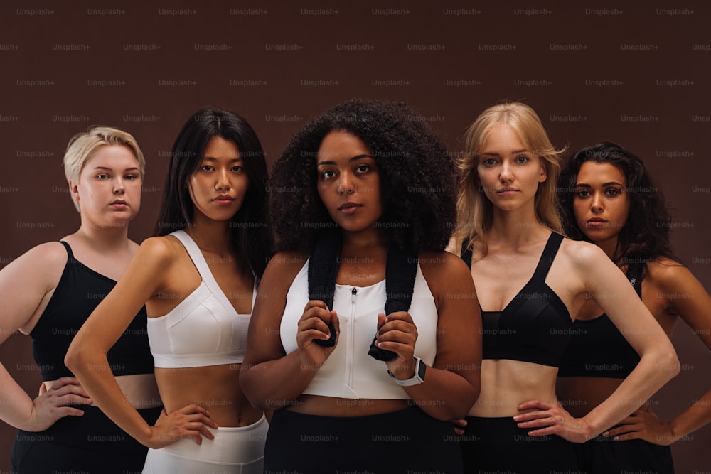 Diverse Frauen in Sportbekleidung schauen in die Kamera, während sie vor einem braunen Hintergrund stehen. Fünf Weibchen verschiedener Rassen im Studio.