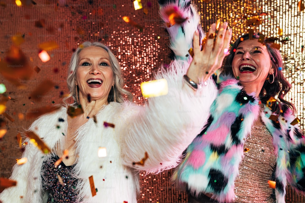 Zwei fröhliche Seniorinnen in Pelzmänteln tanzen unter Konfetti vor Glitzerkulisse