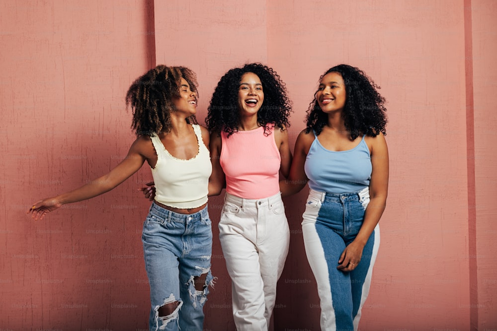Three happy women with curly hair walk together. Young females having fun at a pink wall.