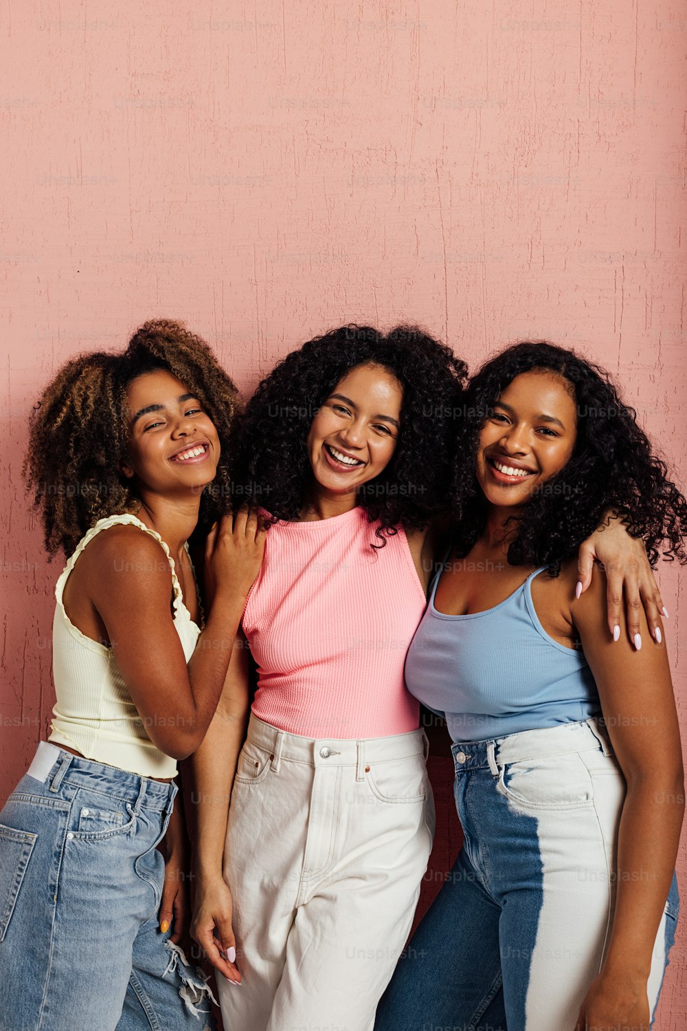 Trois femmes souriantes aux cheveux bouclés se tenant ensemble et regardant la caméra contre un mur rose