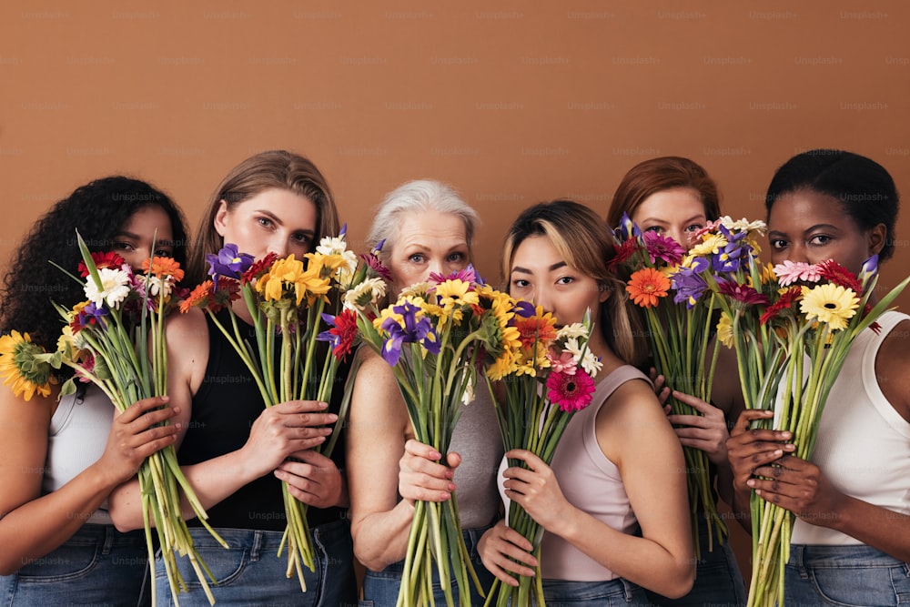 Group of diverse women hide their faces by bouquets of flowers. Portrait of six females of a different race, age, and figure type.