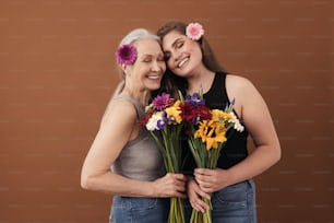 Due donne sorridenti di età diverse in piedi testa a testa in studio. Femmine caucasiche con mazzi di fiori in mano e fiori tra i capelli.