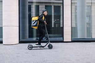 Cheerful female courier standing on electric scooter. Woman in cycling helmet wearing thermal backpack looking away.