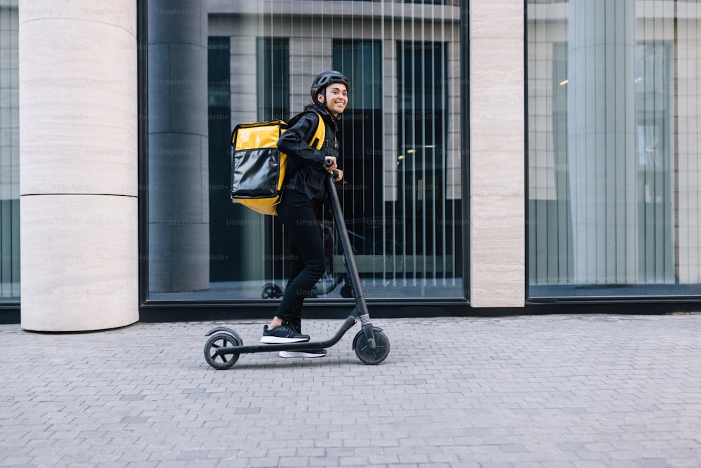 Cheerful female courier standing on electric scooter. Woman in cycling helmet wearing thermal backpack looking away.