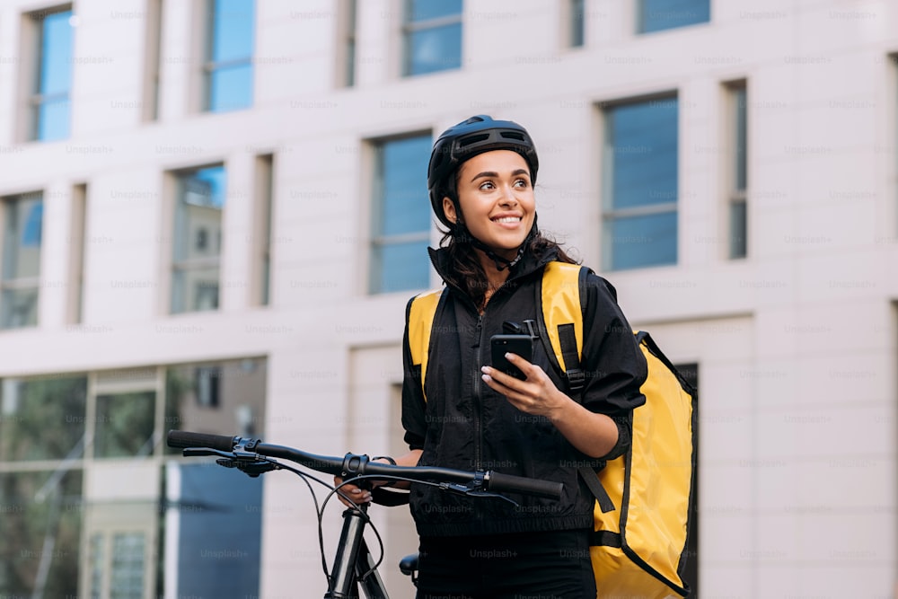 Donna di consegna del cibo che utilizza lo smartphone sulla strada in città. Corriere donna con casco da ciclismo con zaino che distoglie lo sguardo.