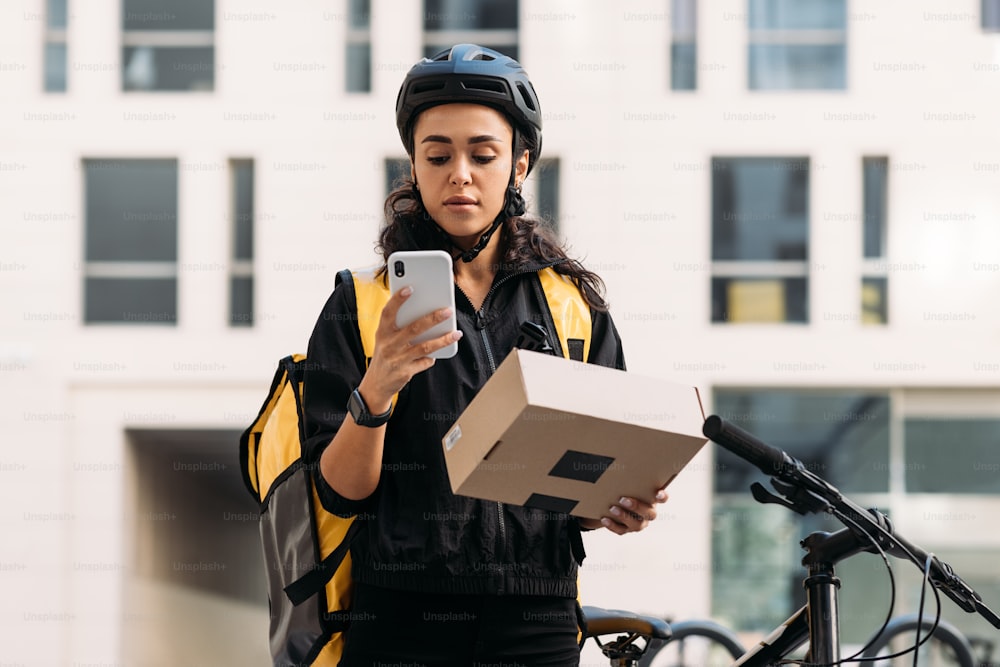 Donna di consegna in casco da ciclismo che tiene una scatola di cartone che controlla le informazioni sul telefono cellulare