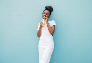 Mujer joven vestida de blanco bebiendo jugo de naranja mientras está parada en la pared azul