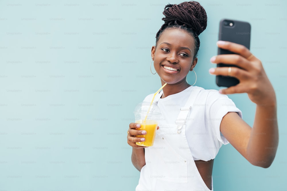 Joven mujer sonriente sosteniendo un jugo y tomándose una selfie mientras está parada al aire libre en la pared azul