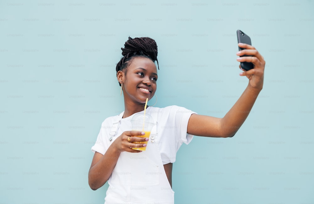 Happy woman drinking juice and taking selfie near a blue wall outdoors
