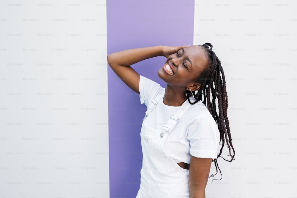 Mujer feliz con los ojos cerrados con ropa blanca apoyada en la pared blanca con rayas púrpuras