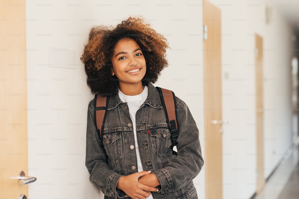 Young student with backpack smiling in high school leaning a wall