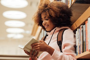 Menina em casuais com mochila inclinada estante na biblioteca. Aluno lendo um livro enquanto está na biblioteca.