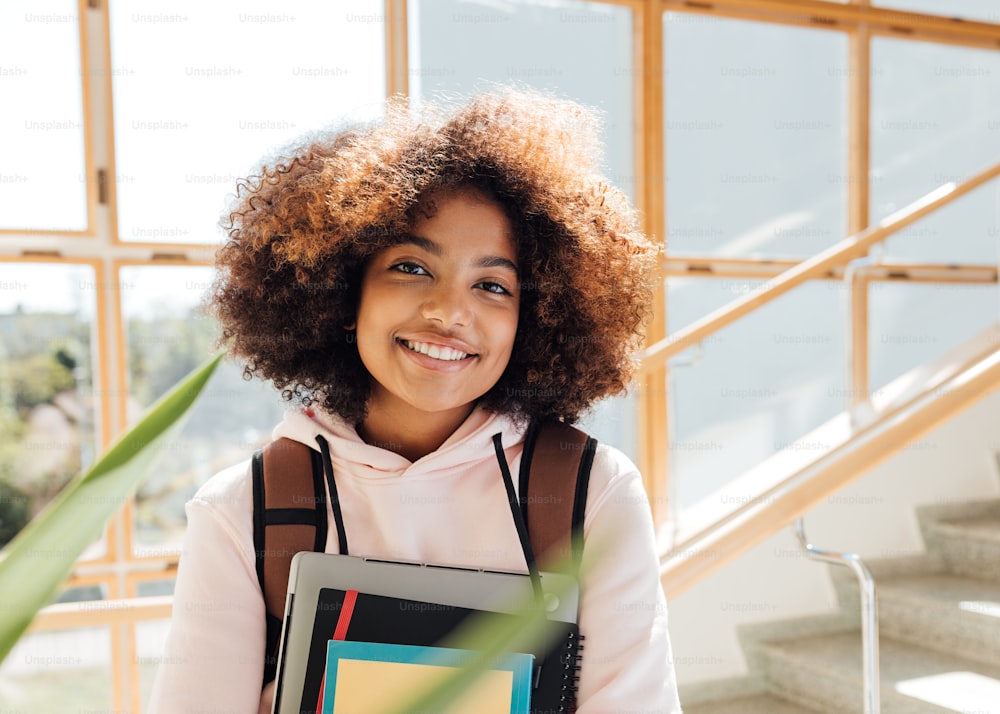 Portrait of a beautiful smiling girl with laptop and books standing in school on stairs and looking at camera