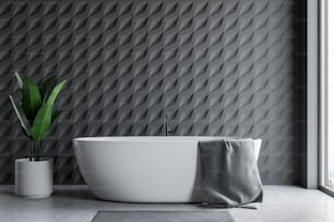 Interior of loft bathroom with gray tile walls, concrete floor, white bathtub with gray towel hanging on it and big potted plant. 3d rendering