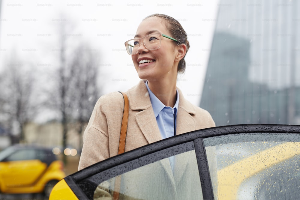 Retrato de una joven y bonita mujer de negocios asiática que sube a un taxi en la lluviosa calle de otoño, sonriendo y mirando hacia otro lado mientras abre la puerta del coche