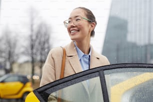 Portrait of young pretty  Asian businesswoman getting in  taxi cab on rainy autumn street, smiling and looking away while opening car door