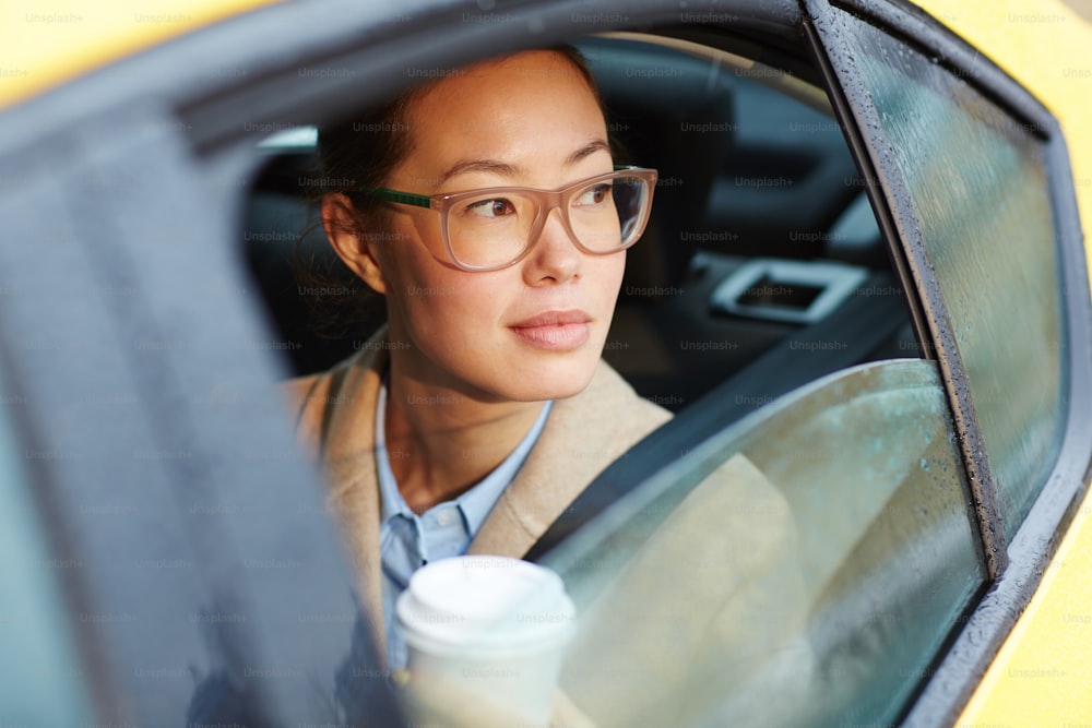 Retrato da empresária asiática confiante que anda no banco de trás do carro olhando para fora da janela iluminada pela luz do sol, segurando a xícara de café