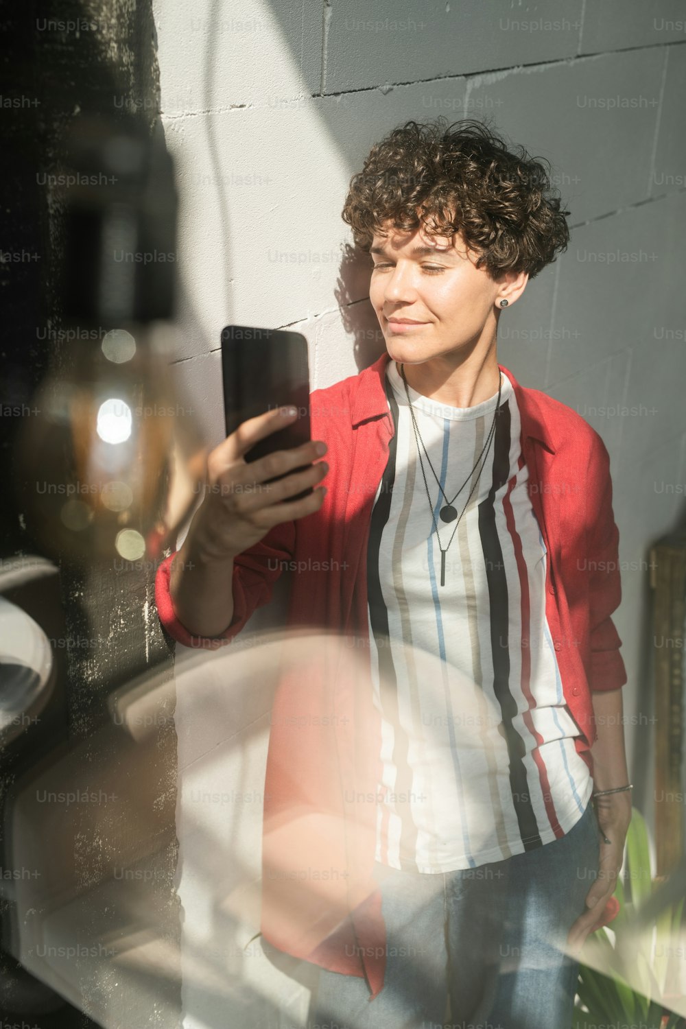 Elegant young woman with mobile gadget standing by wall and making selfie while looking at camera in smartphone