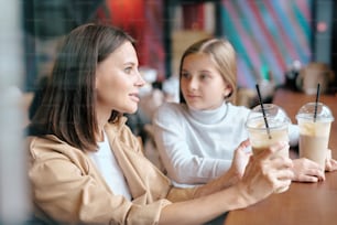 우유 칵테일을 마시며 귀여운 딸과 이야기를 나누는 예쁜 젊은 여성이 카페에서 테이블 옆에 앉아 술을 마시고 있다