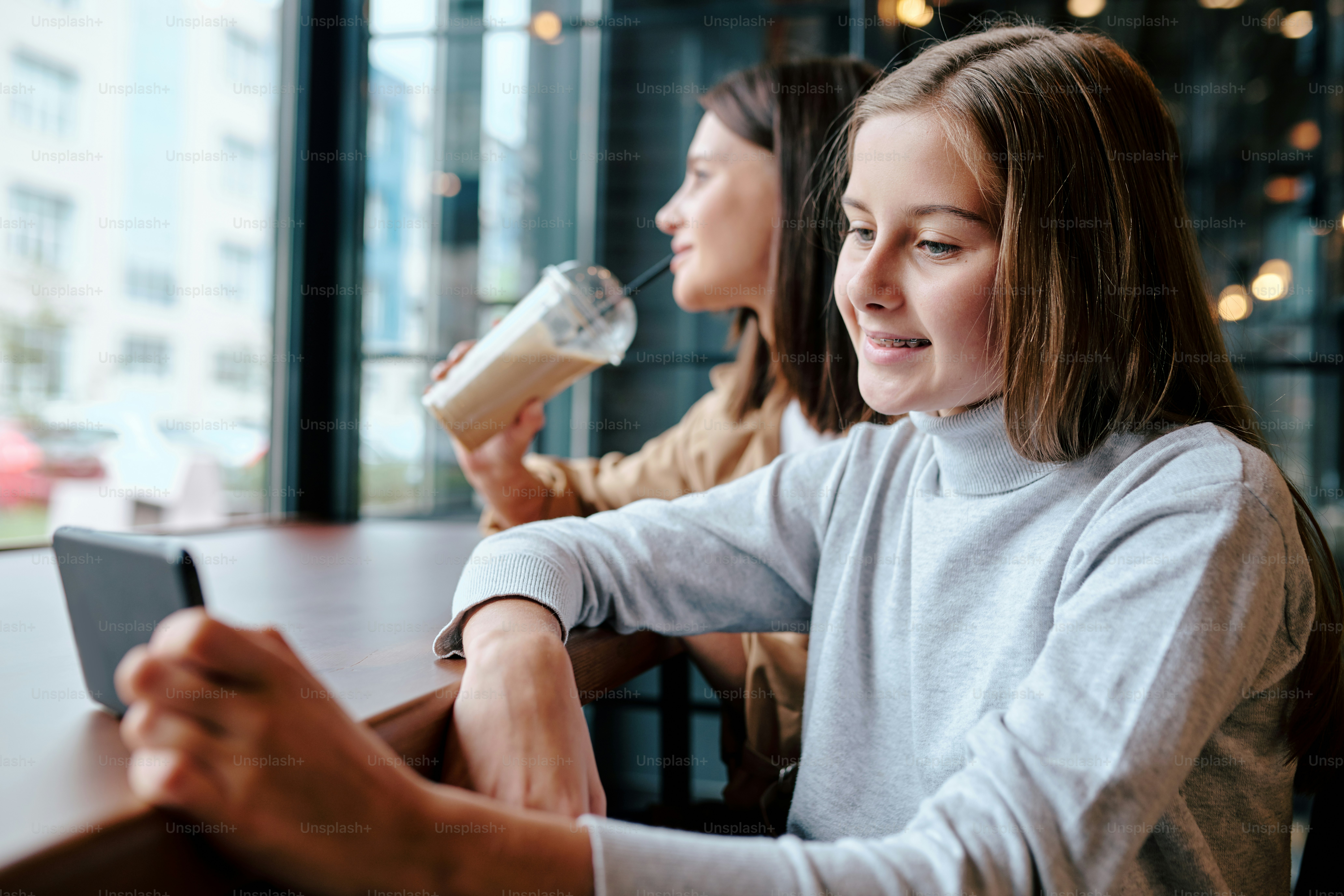 Smiling girl with smartphone sitting by table in cafe and watching video with her mother having cocktail on background