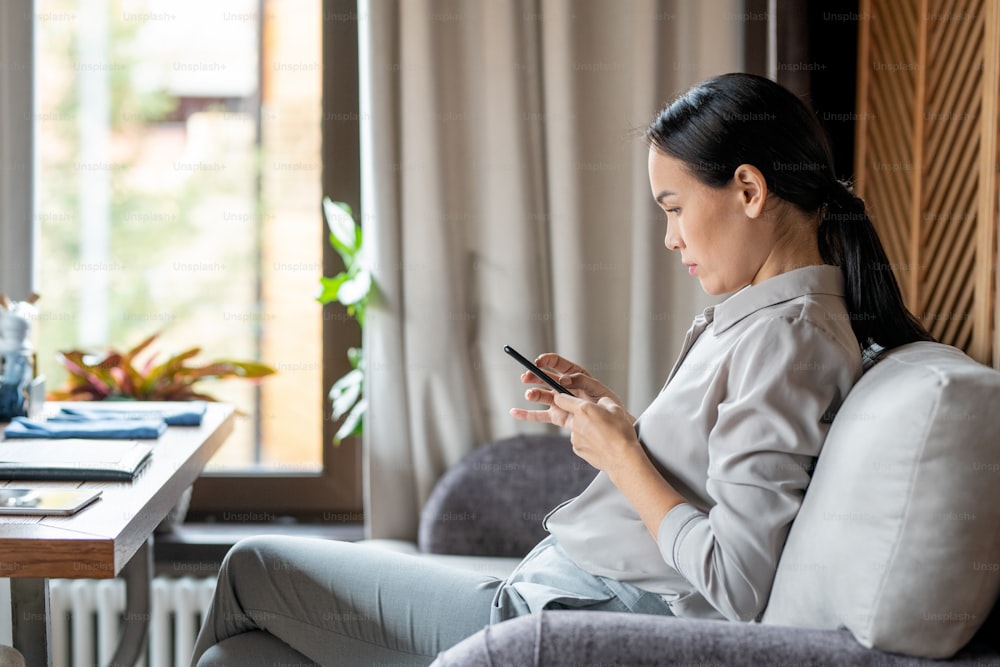Mujer joven casual con teléfono inteligente sentada en el sofá del restaurante y enviando mensajes mientras espera a alguien
