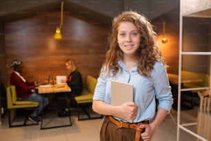 Pretty young smiling student with mobile gadget standing in front of camera in cafe on background of working groupmates