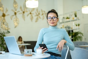 Mulher casual muito jovem com smartphone olhando para você enquanto sentado à mesa na frente da câmera no café