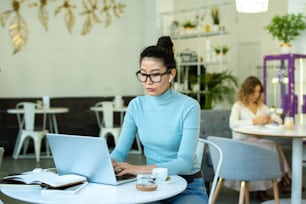 Mujer casual bastante joven con auriculares inalámbricos escribiendo frente a la computadora portátil mientras se conecta en la mesa en la cafetería