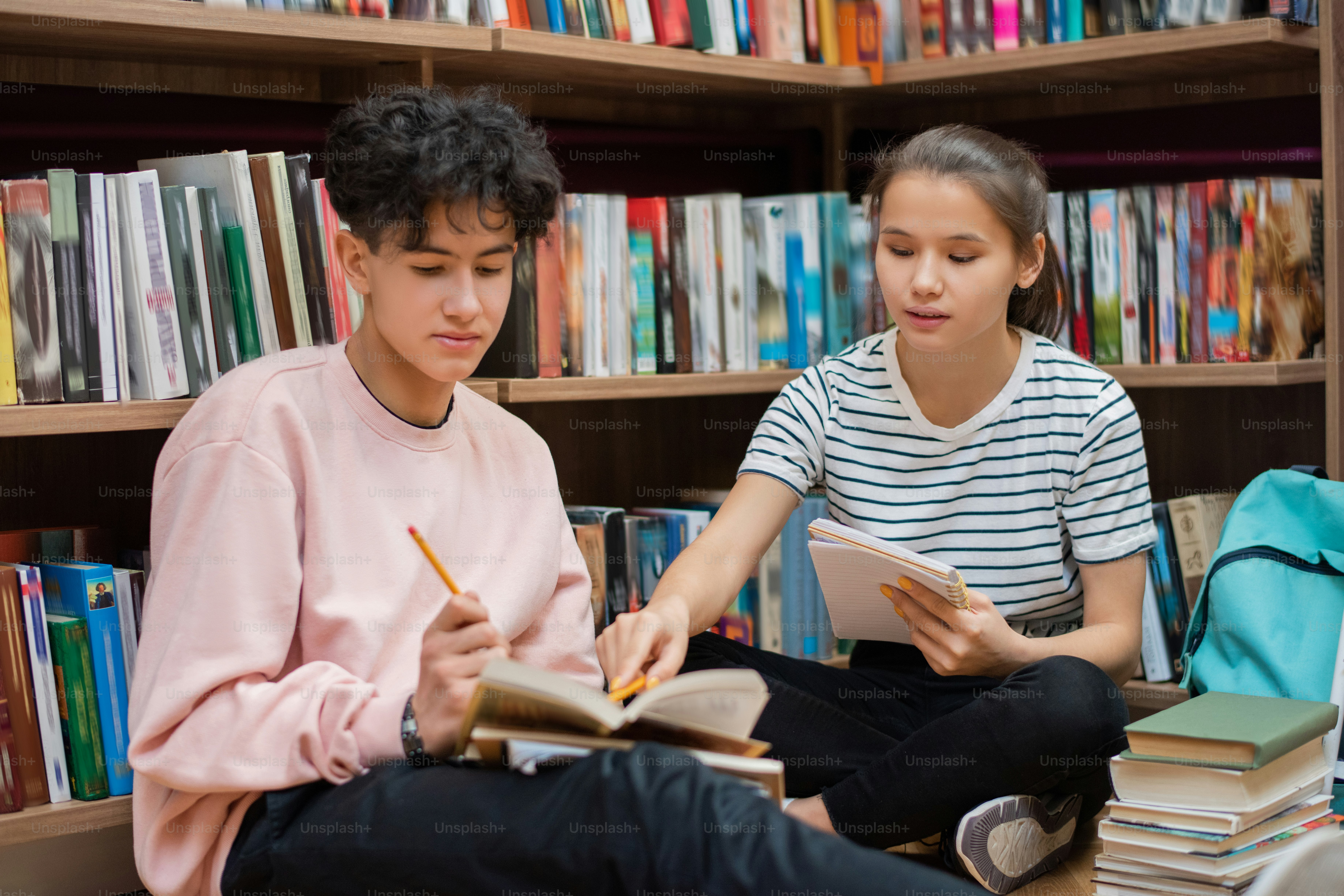 Clever confident girl pointing at page of book held by her classmate during preparation for seminar in college library