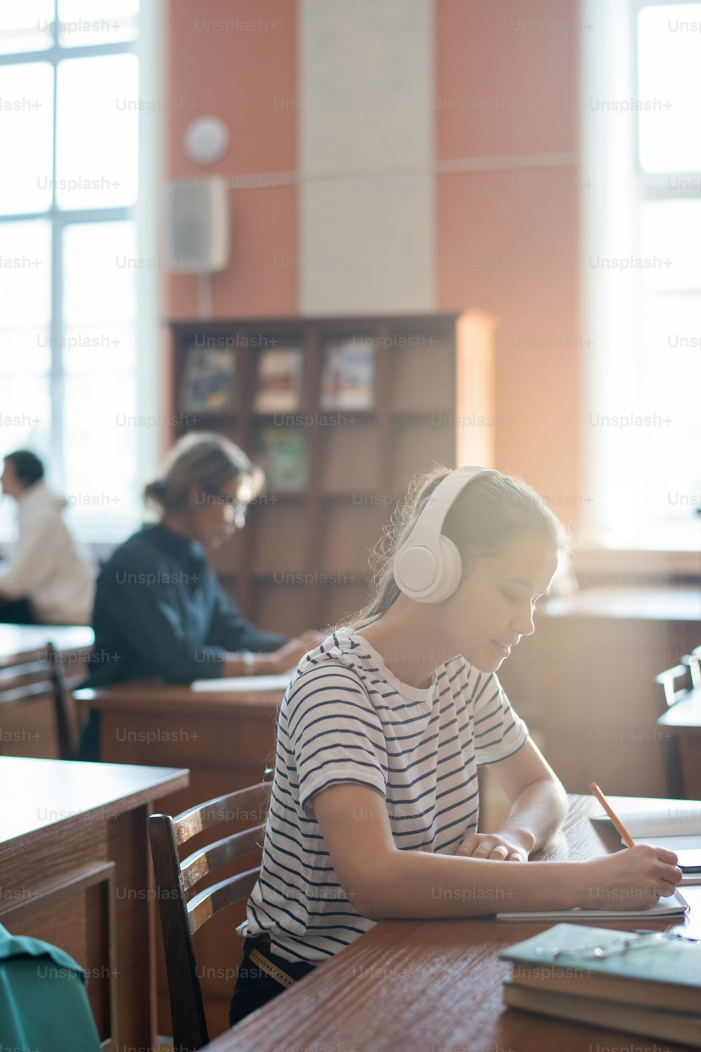 Fleißiges Teenager-Mädchen mit Kopfhörern, das Musik hört und ihre Ideen zu Seminarpunkten in der College-Bibliothek aufschreibt