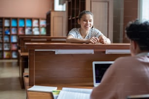 Happy teenage girl looking at her classmate with laptop during conversation in college library after classes