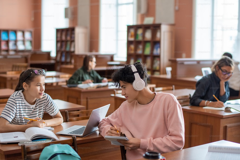 Dos adolescentes discuten puntos del seminario mientras revisan libros y fuentes en línea en la biblioteca de la universidad después de las lecciones
