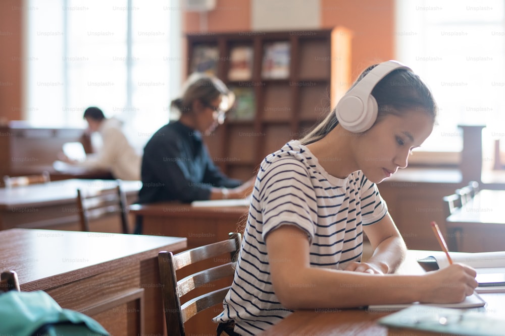 Joven estudiante con auriculares concentrado en la preparación para el seminario mientras toma notas en el escritorio de la biblioteca de la universidad
