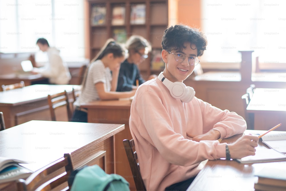 Adolescente sonriente con anteojos y ropa casual mirándote mientras está sentado junto al escritorio en la biblioteca de la universidad y estudiando