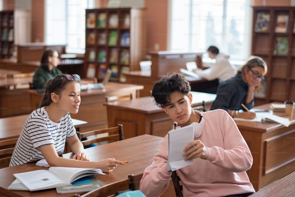 10대 남자가 쉬는 시간에 대학 도서관에서 세미나 계획에 대해 토론하는 동안 동급생에게 메모장에 노트를 보여주고 있다