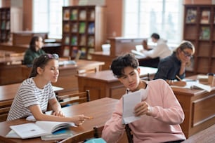 Teenage guy showing his notes in notepad to classmate during discussion of seminar plan in college library at break