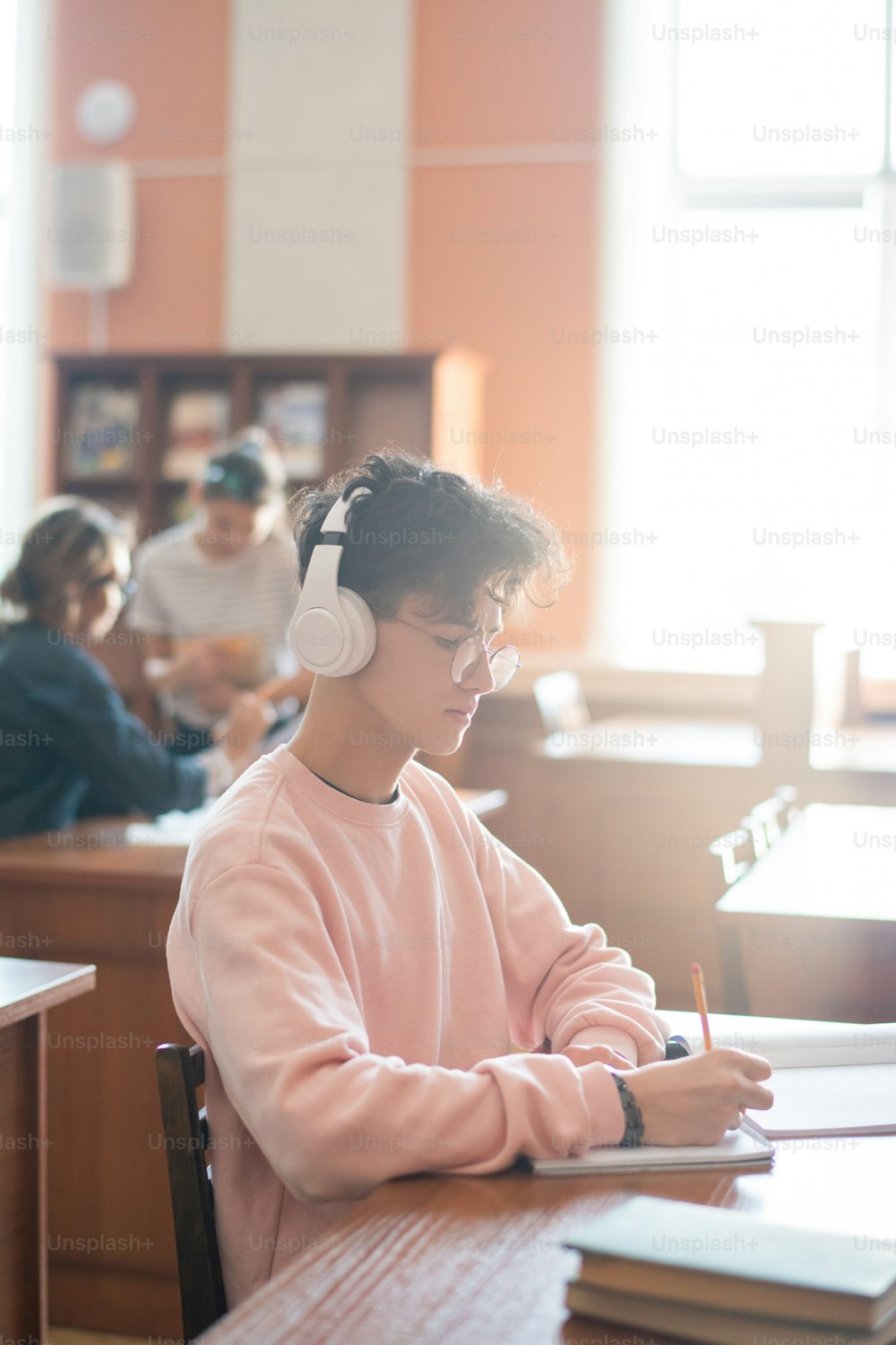 Serious student with headphones concentrating on making plan of report while making notes in notepad in college library